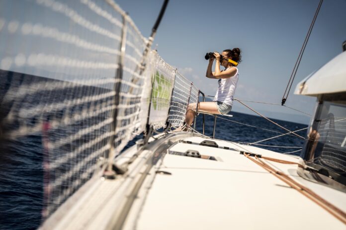 una donna seduta a bordo di una barca, dietro il parapetto fatto di rete, tiene in mano un binocolo
