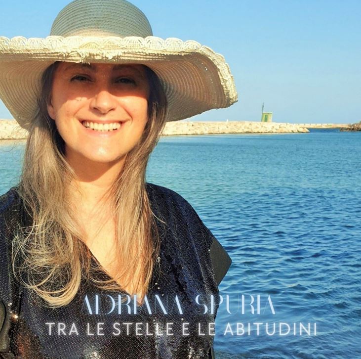 tra le stelle e le abitudini - adriana spuria in primo piano, sorridente, indossa un prendisole nero e un cappello di paglia bianco. Sullo sfondo il mare