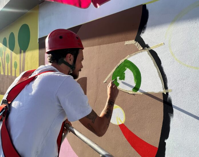 GeometricBang con casco protettivo e cinture di sicurezza sta dipingendo la facciata del palazzo realizzando la sua opera con colori verde in un cerchio beige