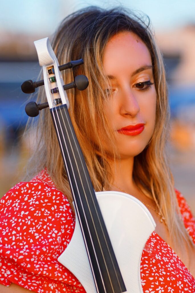 beatrice limonti indossa un abito estivo di cotone a fiori, e ha un violino bianco appoggiato alla spalla destra