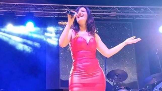 laura dimitri in primo piano durante un concerto, indossa un vestito rosso aderente e ha il microfono nella mano destra