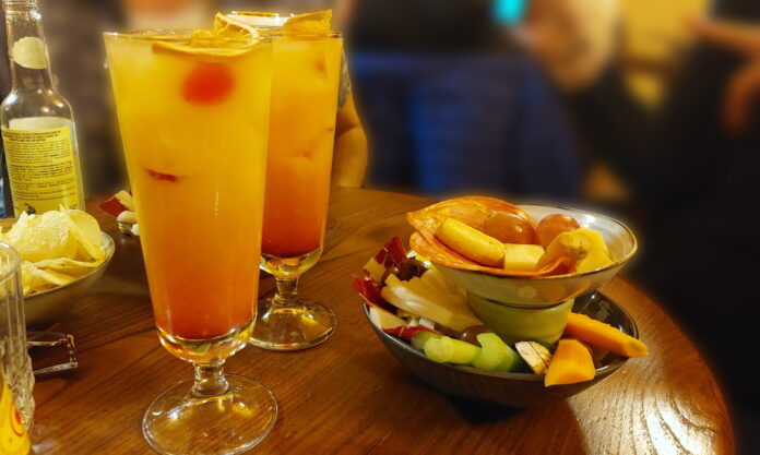 Aperitivo happy hour - nella foto due bicchieri a cono con dentro un cocktail giallo e ciliegine. I bicchieri sono appoggiati su un tavolo rotondo di legno e sul tavolo ci sono anche delle ciotole con dentro degli stuzzichini