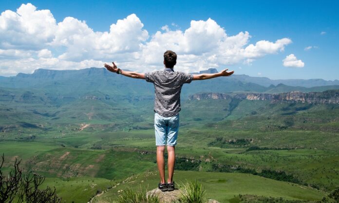 Green Communities - un uomo in cima ad un masso su una montagna è di schiena, ha le braccia aperte ed è rivolto verso la vallata. Insdossa un bermuda di jeans e una maglietta grigia