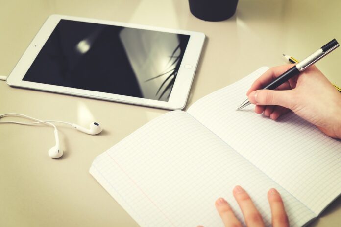 una donna sta scrivendo su un notes e vicino c'è un tablet con degli auricolari