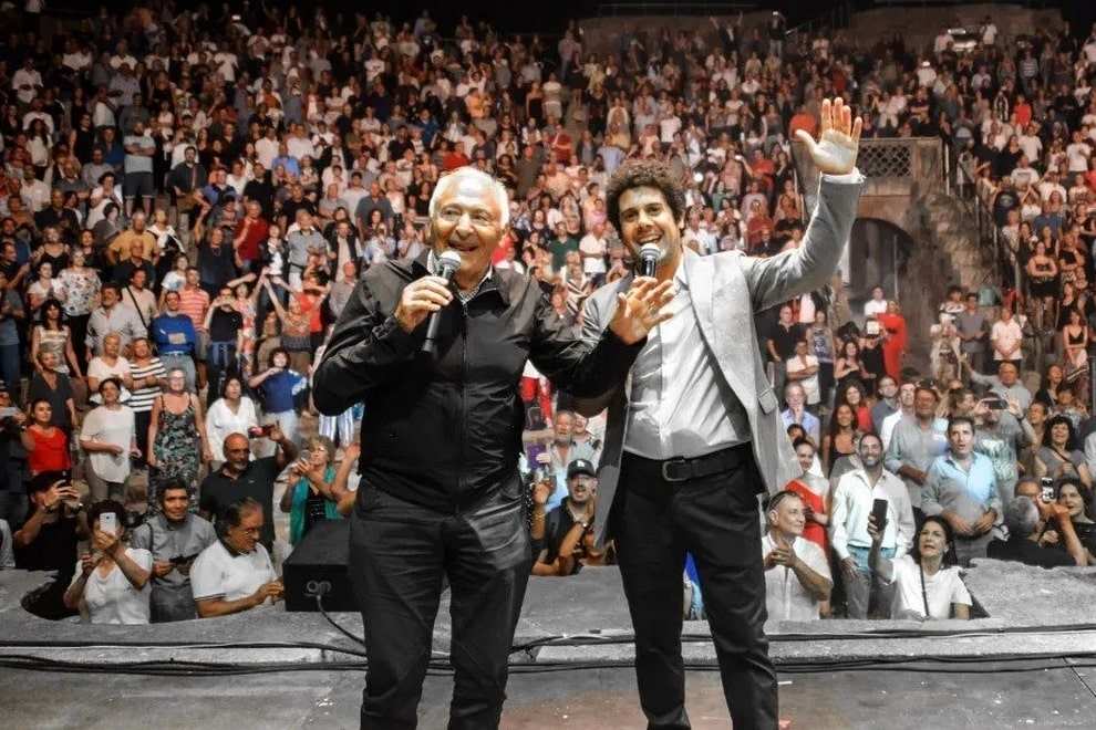 mogol (a sinistra) e gianmarco carroccia (a destra) insieme sul palco, con il pubblico alle spalle