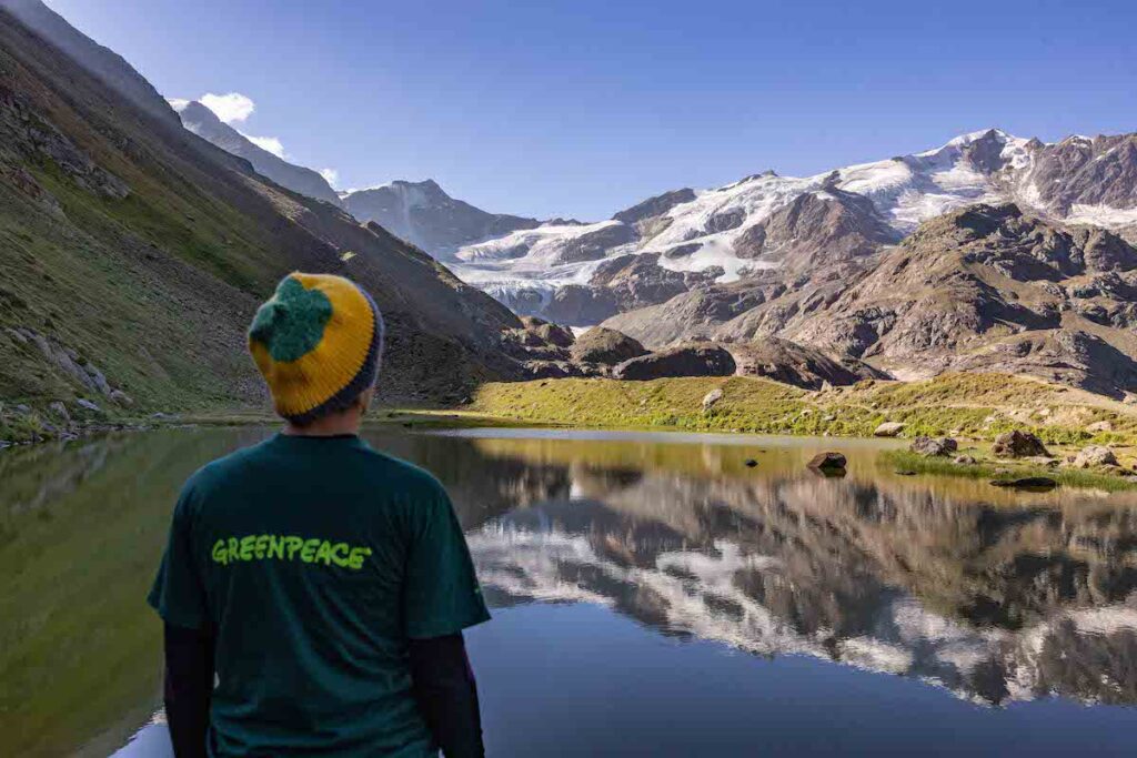 un uomo di schiena con un maglione verde con la scritta "Greenpeace" e berretto di lana, sta guardando un lago e una montagna 