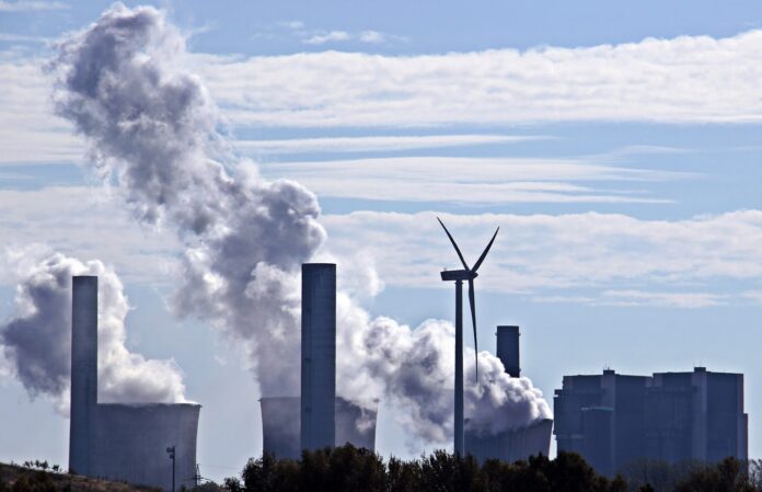 energia e greenwashing - nella foto una centrale elettrica con ciminiere, una pala eolica e tanto fumo