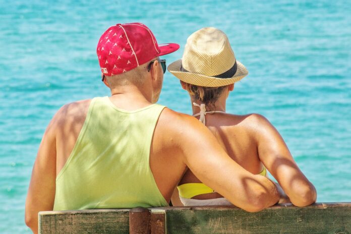 over 50 - una coppia, uomo e donna cinquantenni, di schiena guardano il mare. Lui indossa una canottiera gialla e un capellino rosso, ha i capelli rasati. Lei indossa un top e un cappellino di paglia ed ha i capelli raccolti