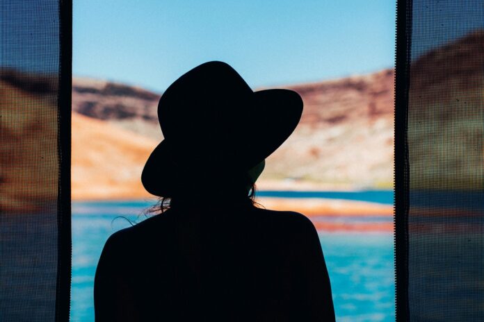 Depressione da Rientro delle Vacanze: Consigli e Strategie - una donna in ombra con cappello a falde larghe è di schiena e sta guardando il mare dalla finestra aperta