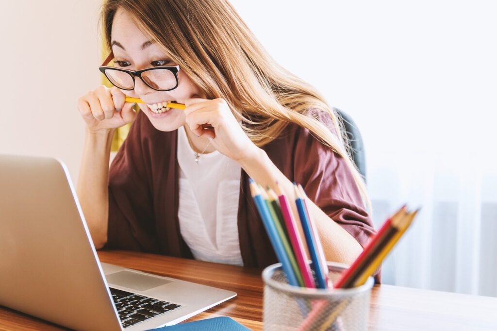 rientro scolastico anno accademico 2023 - una studentessa con capelli lunghi biondi è davanti a un computer e sta stringendo con i denti una matita. Indossa grossi occhiali da vista