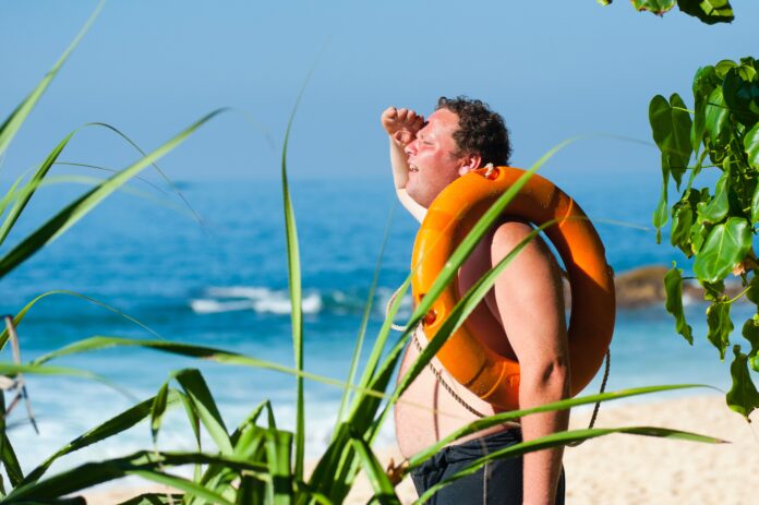 scottature solari - nella foto un uomo panciuto ha un salvagente a tracolla, indossa un acanottiera bianca, con la mano destra si copre gli occhi dal sole, è su un aspiaggia e si vde il mare e l'umo è visibilmente arrossato dal sole