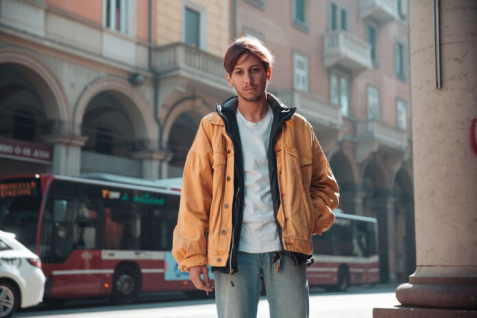 edodacato, fotografato sotto dei portici, indossa jeans, t shirt bianca e un giaccone arancione