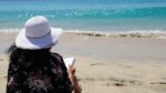 letture estive - una donna è seduta, di schiena all'obiettivo, su una spiaggia e sta leggendo un libro rivolta verso il mare. In dossa un cappello a falde larghe bianco e ha lunghi capelli neri