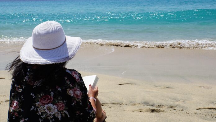 letture estive - una donna è seduta, di schiena all'obiettivo, su una spiaggia e sta leggendo un libro rivolta verso il mare. In dossa un cappello a falde larghe bianco e ha lunghi capelli neri