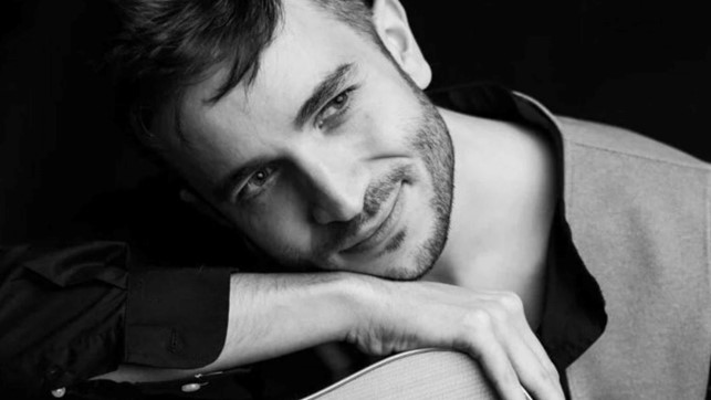 Riccardo D’Avino in una foto in bianco e nero, ha il viso appoggiato alla cassa di una chitarra