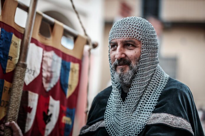 Federico Barbarossa Cocconato d'Asti - foto mezzo busto di un uomo che indossa una maglia d'acciaio medievale sulla testa ed è vestito con abiti d'epoca. Ha barba bianca e sorride, dietro di lui si vede uno stendardo medievale rosso