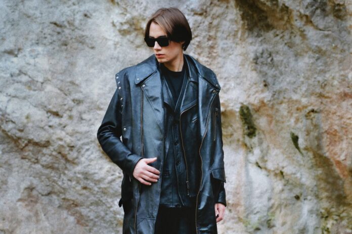 mandark fotogrfto davanti a un muro di pietra, indossa giacca e cappotto di pelle nera