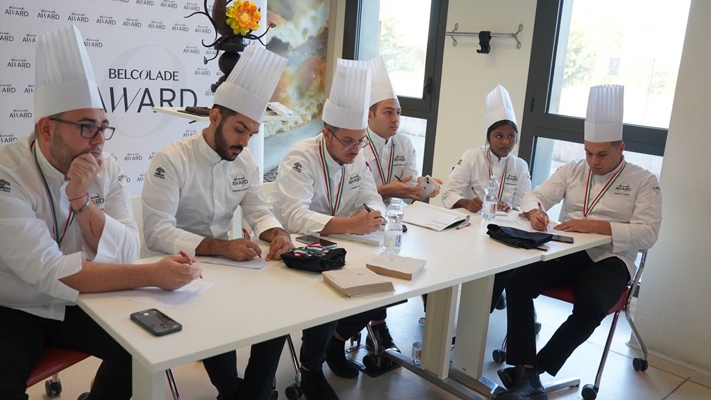 Belcolade award 2023 - i sei finalisti, vestiti con casacca bianca e cappello da chef pasticcere sono seduti ad un tavolo e seguono la lezione