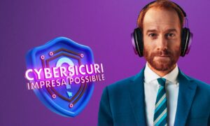 un uomo indossa giacca blu, cravatta a righe blu chiaro e blu scuro e giallo, indossa anche delle cuffie stereofoniche. E' rossiccio di capelli e folta barba rossa. Lo sfondo è color viola e sulla sinistra c'è uno scudo blu e fucsia con la scritta "Cybersicuri - impresa possibile"