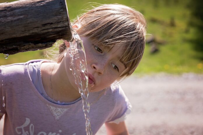 Una bambina con capelli biondi raccolti in una coda e frangetta, indossa un atshirt lilla e sta bevendo da una fontana fatta di legno