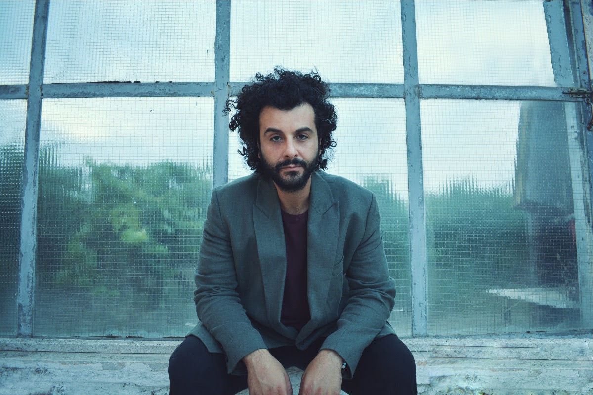 blumosso (barba e capelli neri), è seduto davanti a una finestra, e indossa pantaloni e t-shirt nera e una giacca blu chiaro