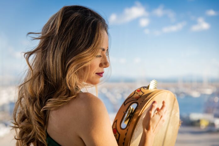 una odnna di profilo tiene in mano un tamburello. Ha lunghi capelli biondi, spalle nude e lo spfndo è un bel panorama di Napoli