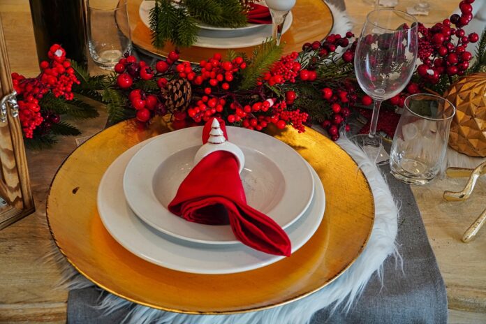 Cenone di Natale - una postazione imbadita a tavola con un piatto con il bordo dorato e sopra altri due piatti, uno piano e uno fondo e dentro un tovagliolo infilato in un segnaposto bianco a forma di albero di natale. Davanti al piatto un ramo di vischio rosso e dei calici