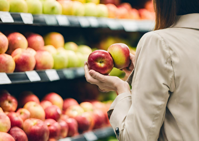 due mele rosse in mano a una donna che è di schiena ed indossa un impermeabile ed è al supermercato di cui si vedono gli scaffali pieni di frutta