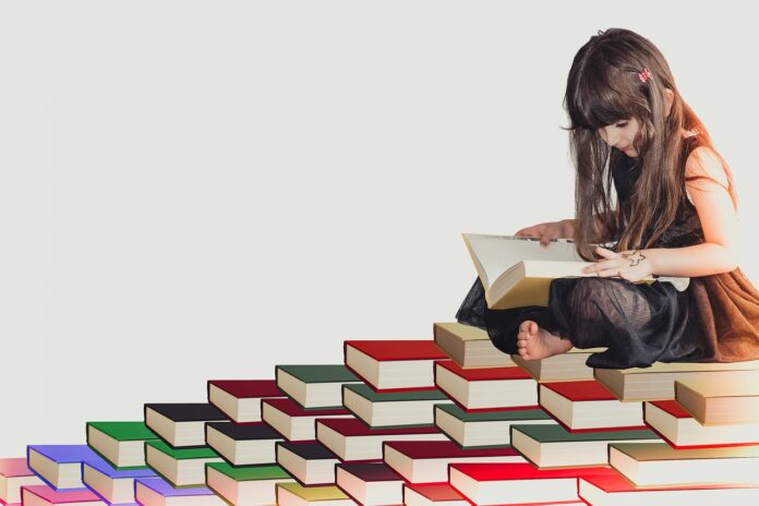 Dei libri messi in modo da formare dei gradini e in cima c'è una bambina seduta con le gambe incrociate che legge. La bambina ha i capelli scuri e lunghi