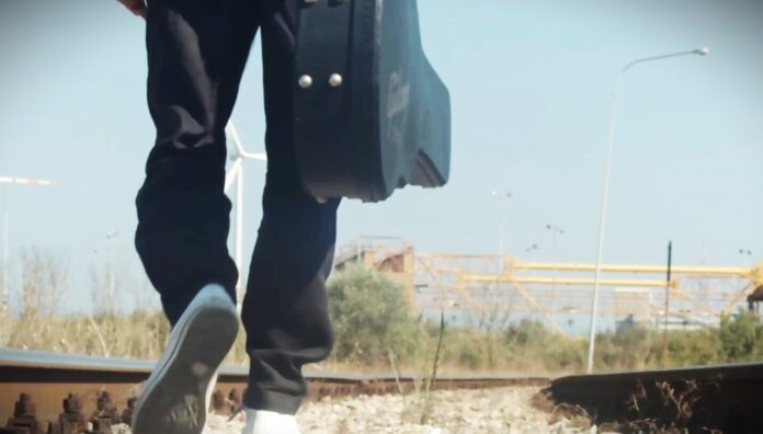 Nicola Ferrari - nella foto si vedono le scarpe e le gambe di un ragazzo mentre cammina di schiena in un astrada sterrata con una custodia di chitarra nella mano destra e introno a lui delle pale eoliche. Indossa dei jeans