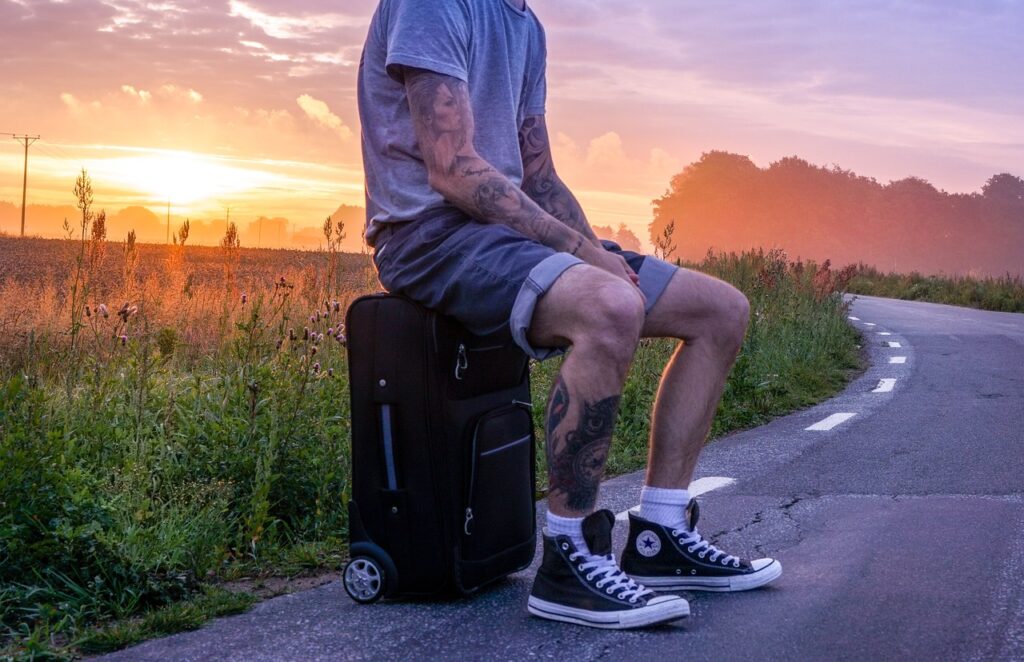 un ragazzo seduto su un avaligia in mezzo a una strada deserta che passa in mezzo all acampagna. Il ragazzo indossa un pantalone corto, una t shirt chiara ed ha tatuaggi sulle braccia e sui polpacci