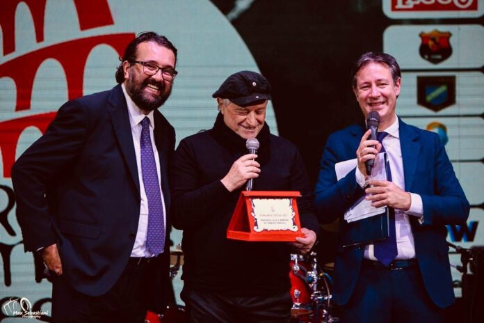 Amedeo MInghi, vestito con giaccone nero, berretto scuro, tiene in mano un microfono e nell'altra una targa di premiazione. E' in mezzo ad altri due uomini, a sinistra Andrea Montemurro e a destra Stefano Raucci