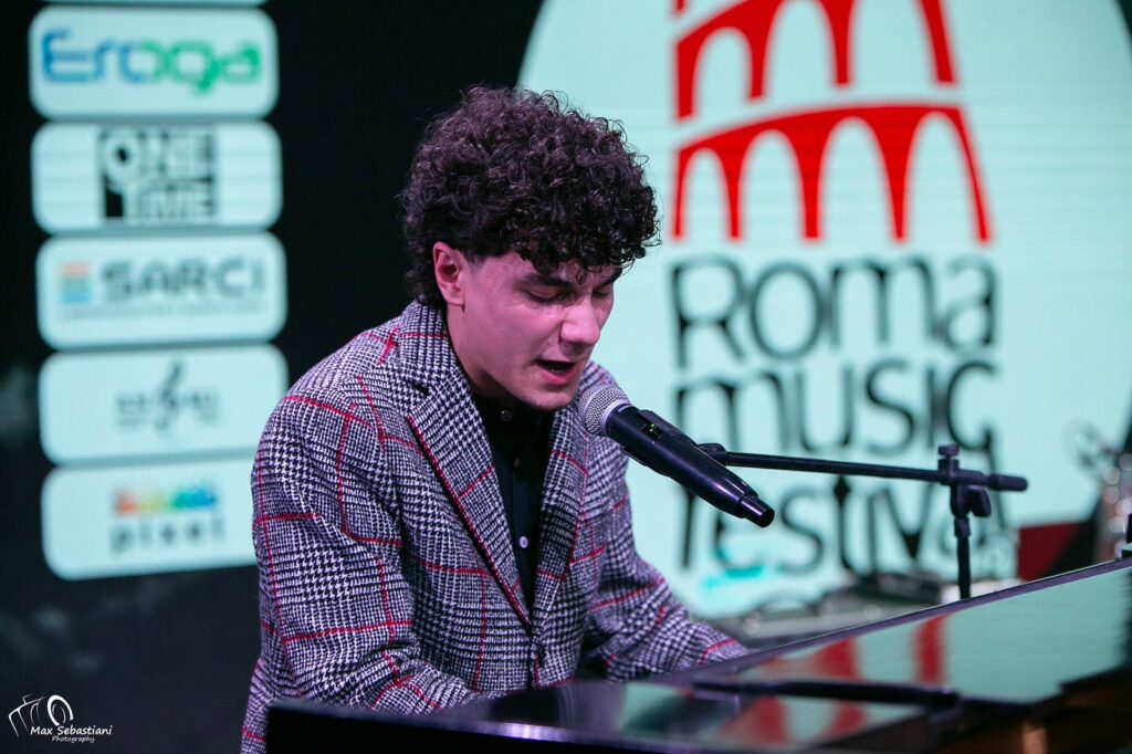 I vincitori del oma Music Festival - un ragazzo con capelli ricci, indossa camicia viola ed è seduto al pianoforte mentre canta