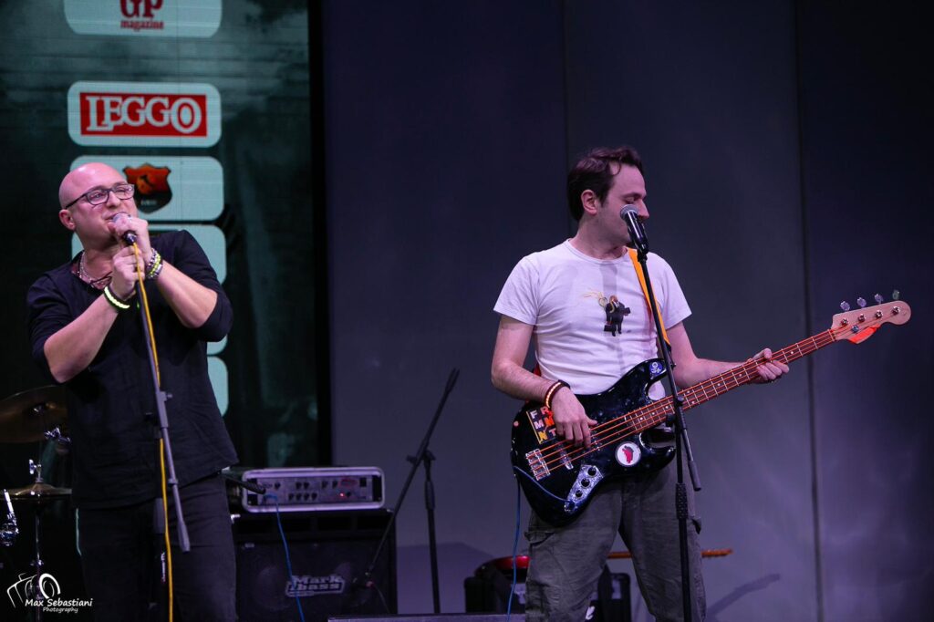 Due ragazzi, uno canta, l'altro suona la chitarra. Il primo indossa abiti scuri, l'altro maglietta bianca e jeans neri