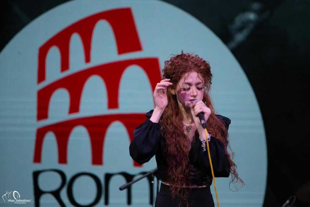 Serata finale Roma Music Festival - una ragazza con midcrofono in mano canta. Ha lunghi capelli rossi ricci  e indossa una giacchetta nera su una camicetta rossa