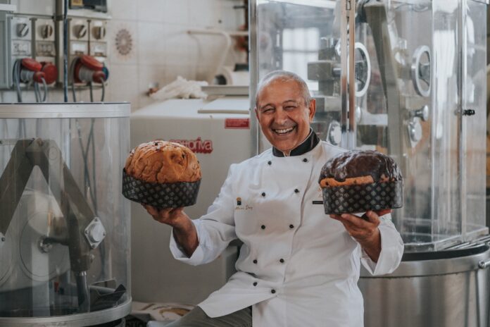 Ferdinando Tammetta seduto tiene in mano due panettoni nel suo lavboratorio di pasticceria dove si vedono dei macchinari per la produzione