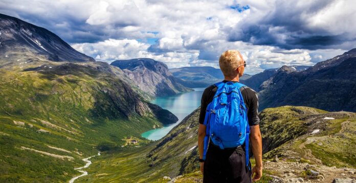 Giornata della montagna - un uomo biondo con zaino blu sulla schiena sta guardando un bellissimo panorama montano con monti e valle con il fiume