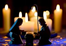 il presepe con sullo sfondo tante candele accese e in primo piano tre figure in ombra, a destra San Giuseppe, a sinistra la Madonna e in mezzo il bamibno gesù. San Giuseppe e Maria sono inchinati sul bambino