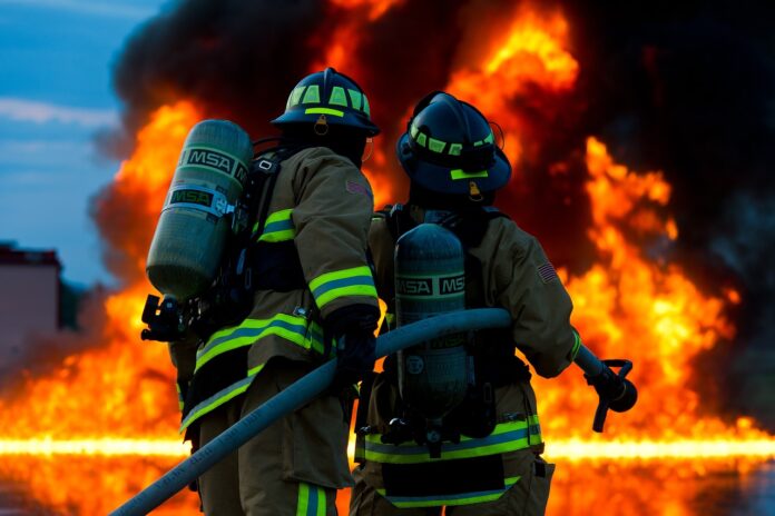 Contaminazione da amianto e PFAS - due vigili del fuoco di schiena, in azione nello spegnimento di un grosso incendio. Indossano tute grige con strisce gialle fluorescenti e bombole