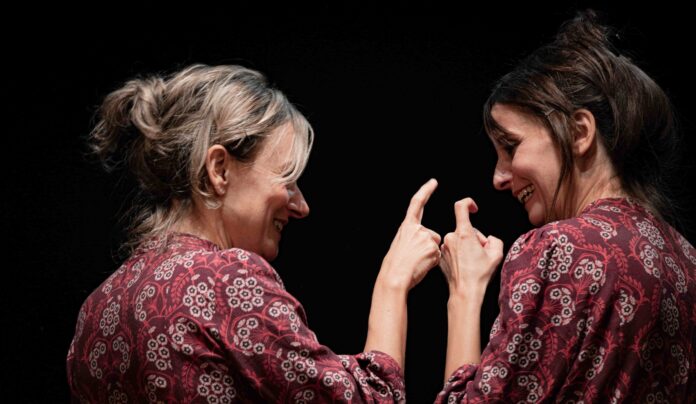 camaleontika - due donne si guardano sorridendo e puntandosi il dito. Vestono lo stesso indumento rosso a fantasia