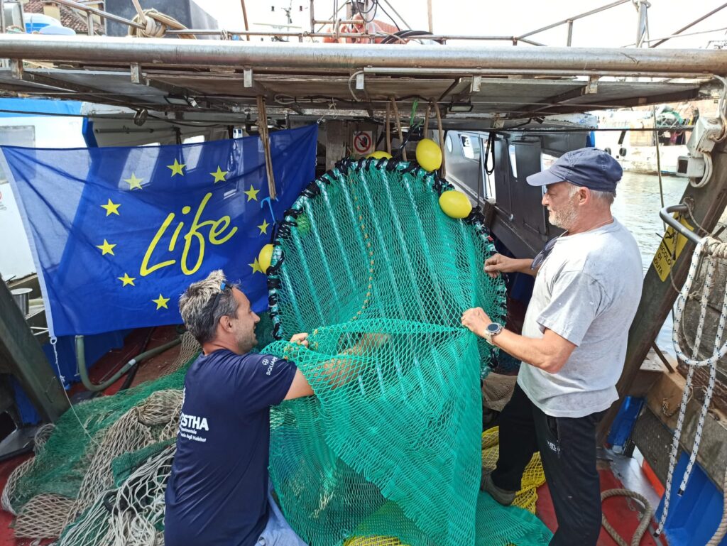 Pesca a strascico - nella foto due uomini, uno con maglietta bianca e cappellino blu, l'altor con maglietta blu, tirano delle reti verdi su una barca dove c'è una bandiera blu con la scritta gialla "life"