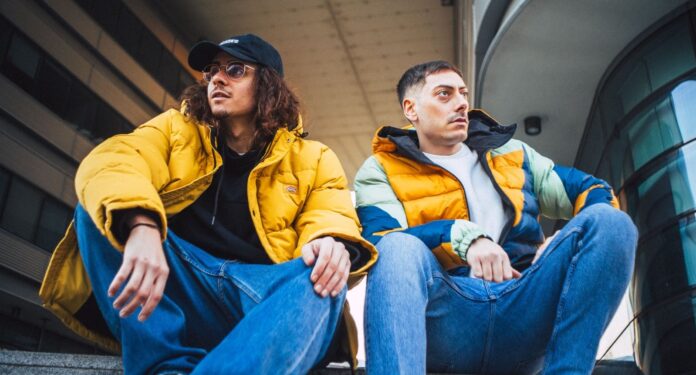 figli di puttana - i liceo paranoia, entrambi seduti, indossano jens azzurri e giacche a vento di colore giallo