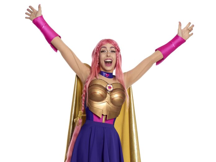 Luli Pampín nella foto una donna con capelli lunghi rosa alza le braccia al cieloe sorride a bocca aperta. indossa guanti lunghi rosa , un corpetto d'oro e una gonna blu, un mantello dorato senza maniche