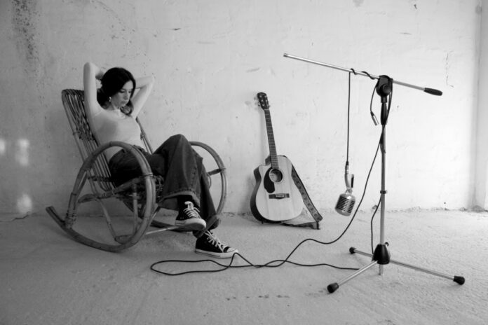 laplastique seduta su una sedia a dondolo, alla sua sinistra una chitarra acustica appoggiata al muro e davanti a lei l'asta di un microfono