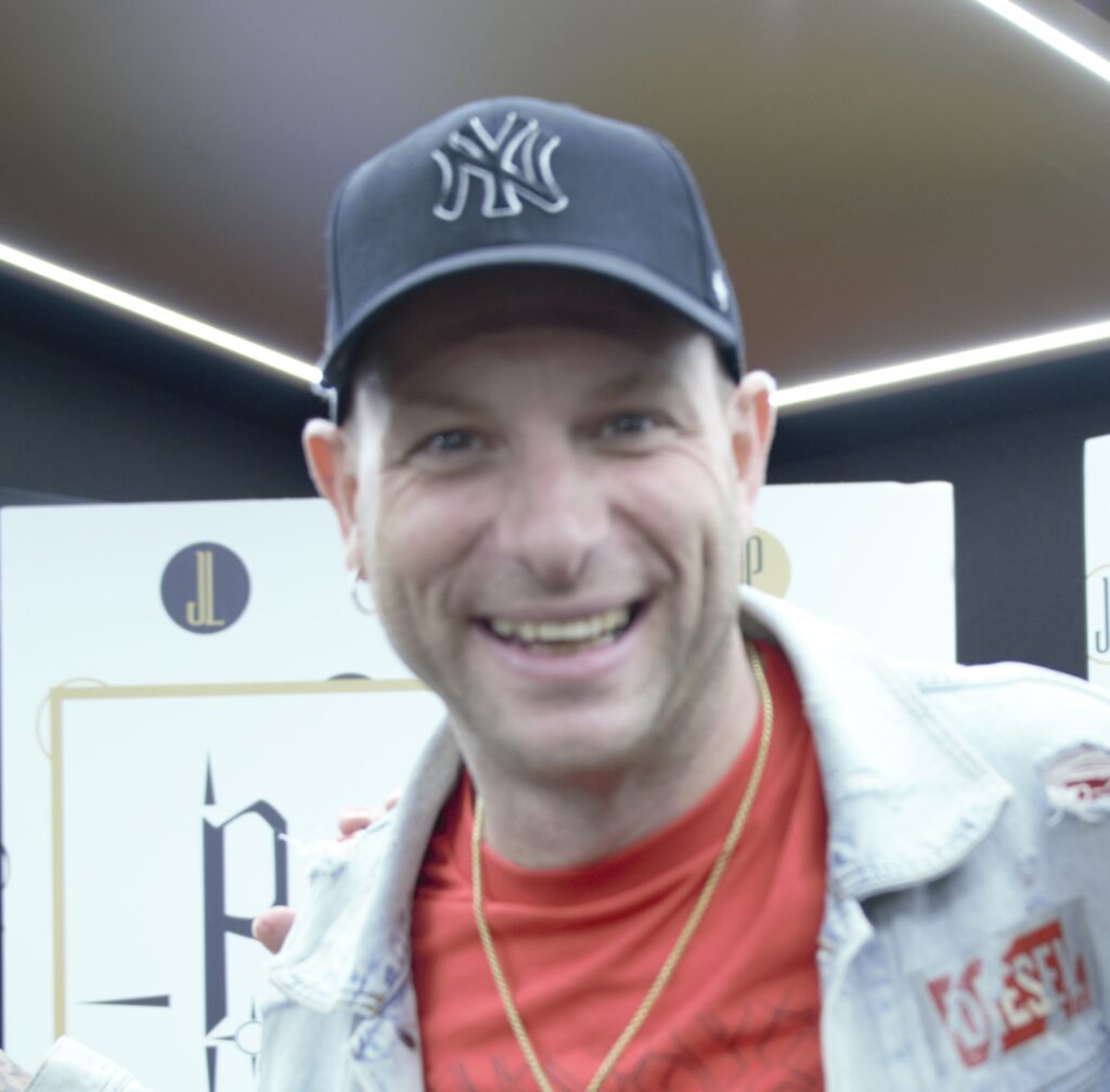 Giani Testa - nella foto., Clementino indosa un cappellino da basket scuro con le lettere NY, un giubbotto di jeans chiaro e una t shirt rossa e sorride