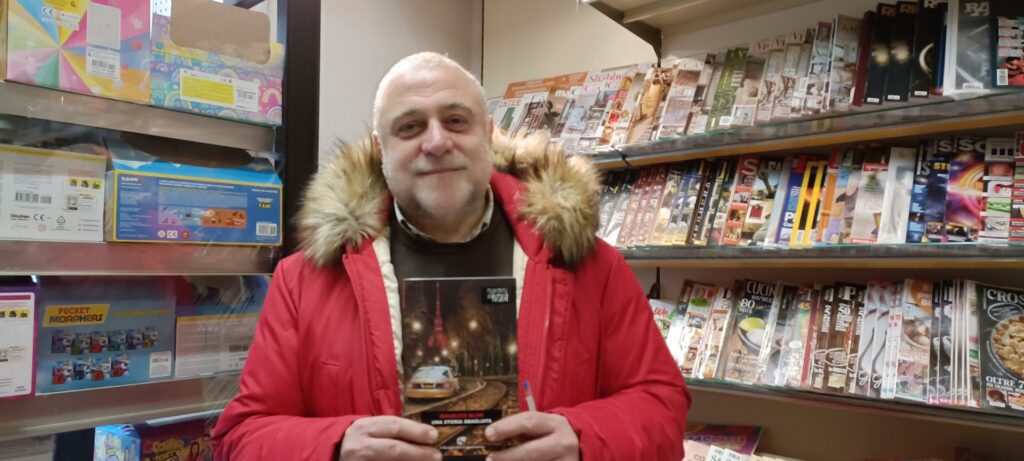 Maurizio Blini con giacca rossa interno libreria con libro in mano