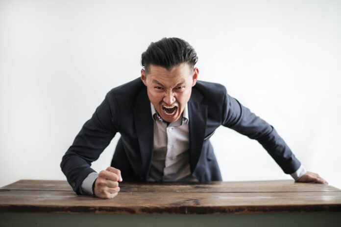 Sindrome di Procuste - un uomo con una mano appoggiata alla scrivania e l'altrachiusa a pugno sta urlando. L'uomo è vestito in giacca e cravatta