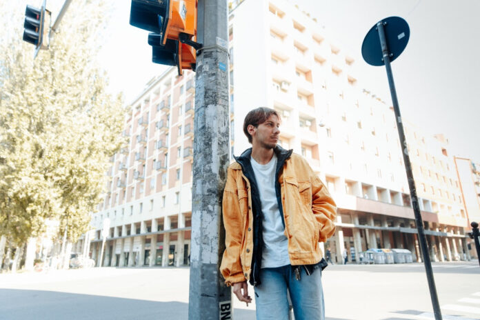 edodacapo fotografato pr strada, appoggiato al palo di un semaforo, indossa jeans azzurri e un giaccone giallo