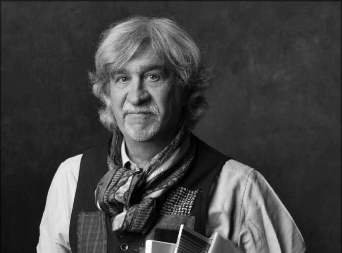 foto in bianco e nero dell'autore del libro che ha lunghi capelli bianchi, barba e baffi, look da rockettaro con camicia bianca, gillet scuro e foulard al collo. tiene sotto il braccio dei dischi di vinile