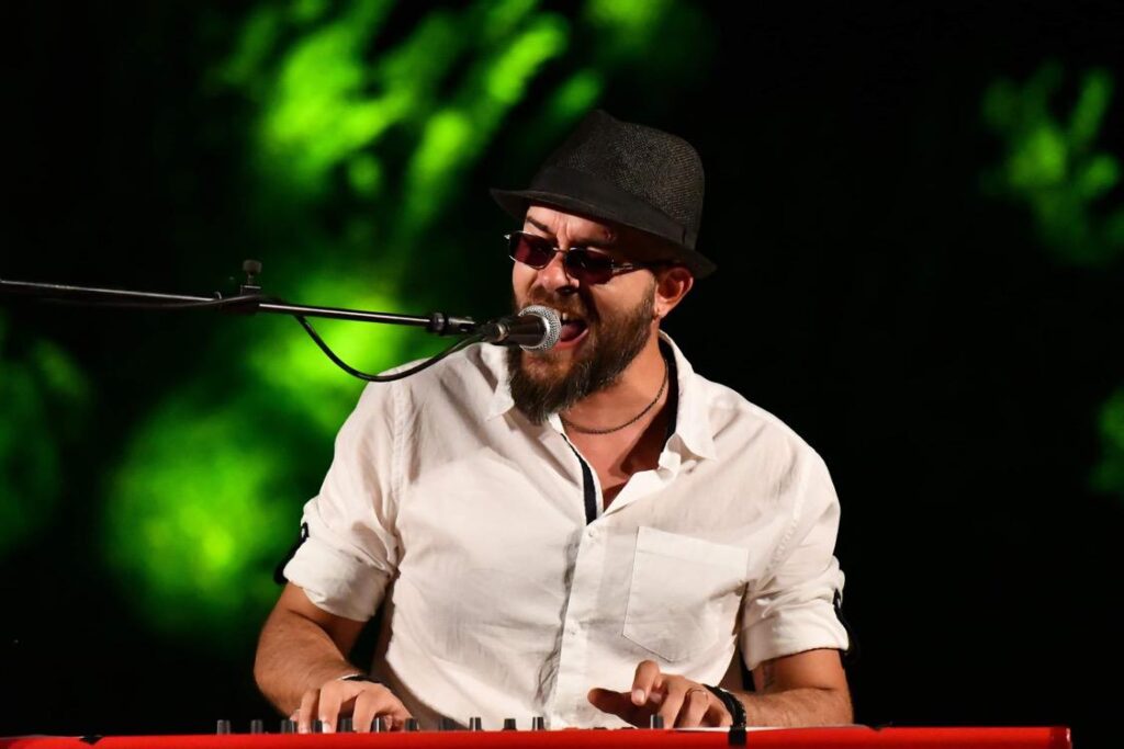 giovani - lorenzo santangelo, camicia bianca, cappello scuro, occhiali da sole e barba, intento a cantare seduoto al pianoforte